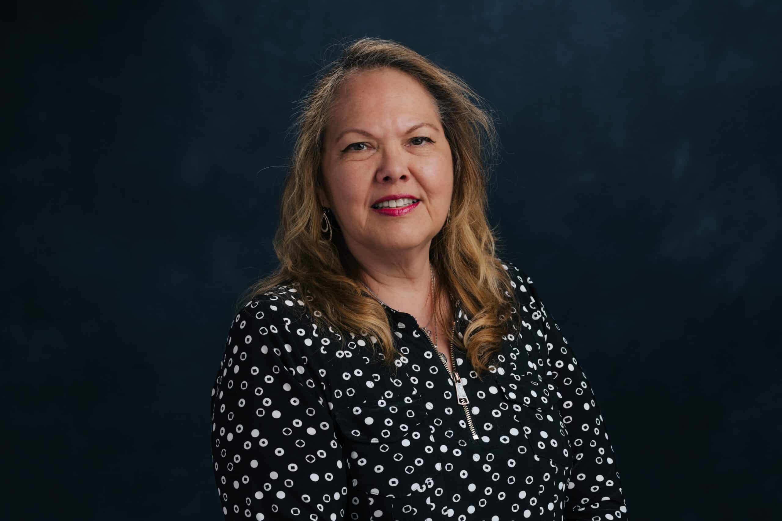 Portrait of a smiling woman with long wavy blonde hair wearing a black polka-dot blouse against a dark backdrop.