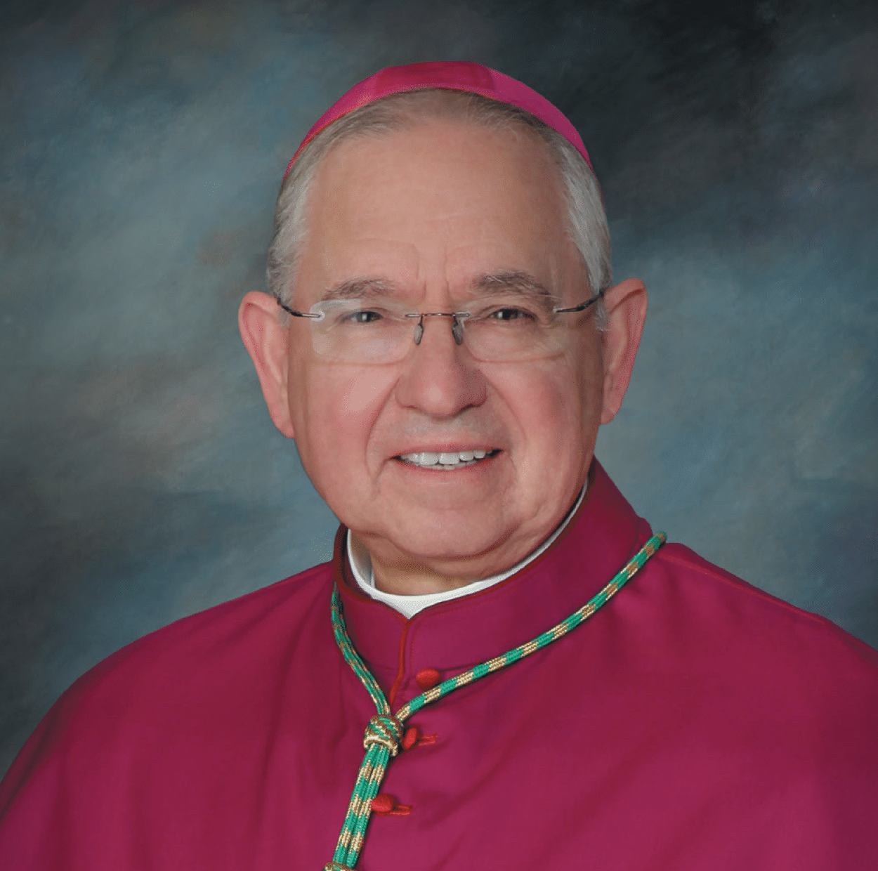 Portrait of a Catholic bishop in magenta liturgical robes with a pectoral cross and glasses on a blue studio backdrop.