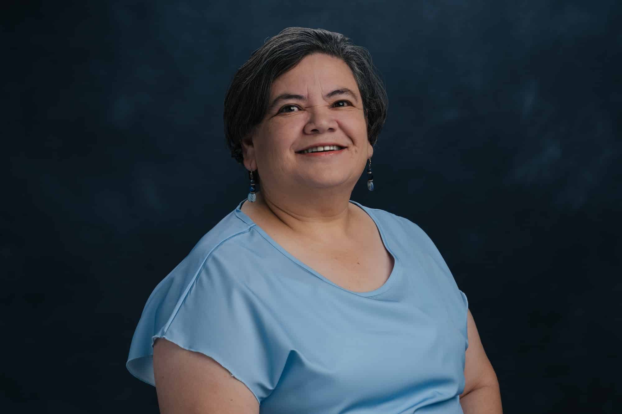 Portrait of a smiling woman with short gray hair wearing a light blue blouse against a dark blue backdrop, earrings visible on both ears.