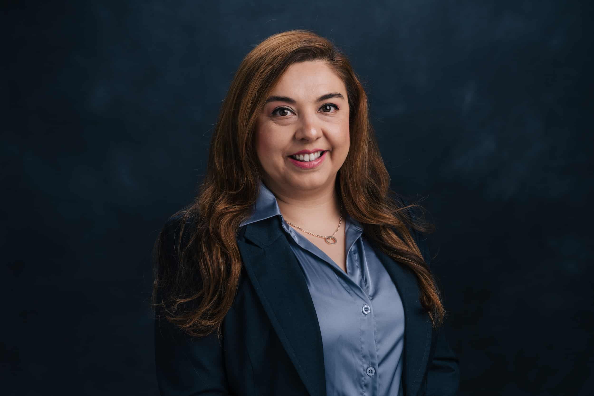 Professional headshot of a smiling woman with long brown hair, wearing a blue blouse and navy blazer against a dark backdrop.