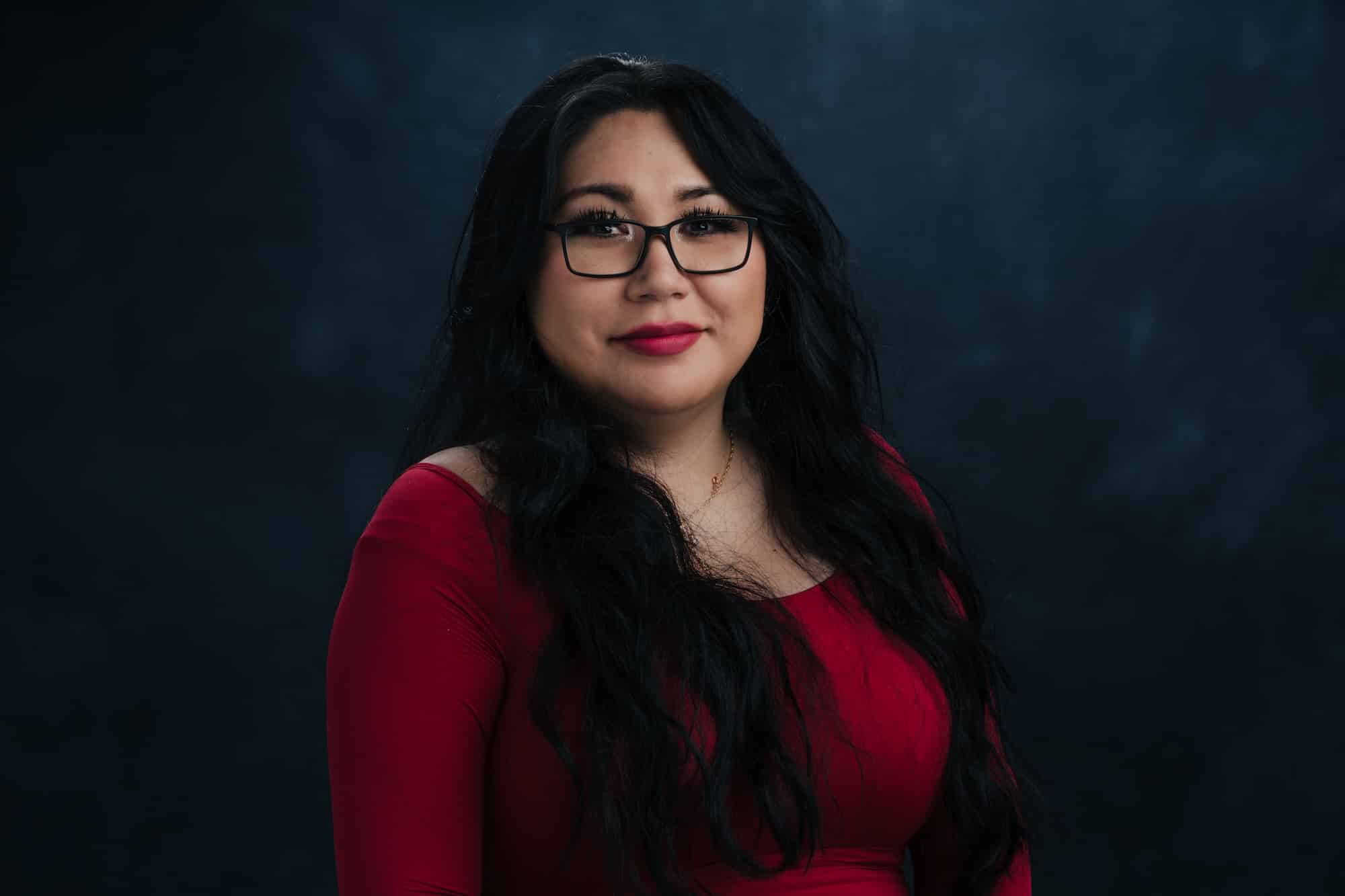 Woman with long dark hair and glasses, wearing a red top, smiling softly against a dark blue backdrop.