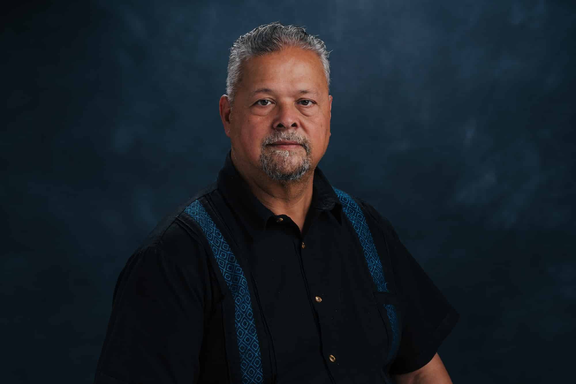 Portrait of a middle-aged man with gray hair and a salt‑and‑pepper goatee, wearing a black shirt with blue suspenders against a dark backdrop.