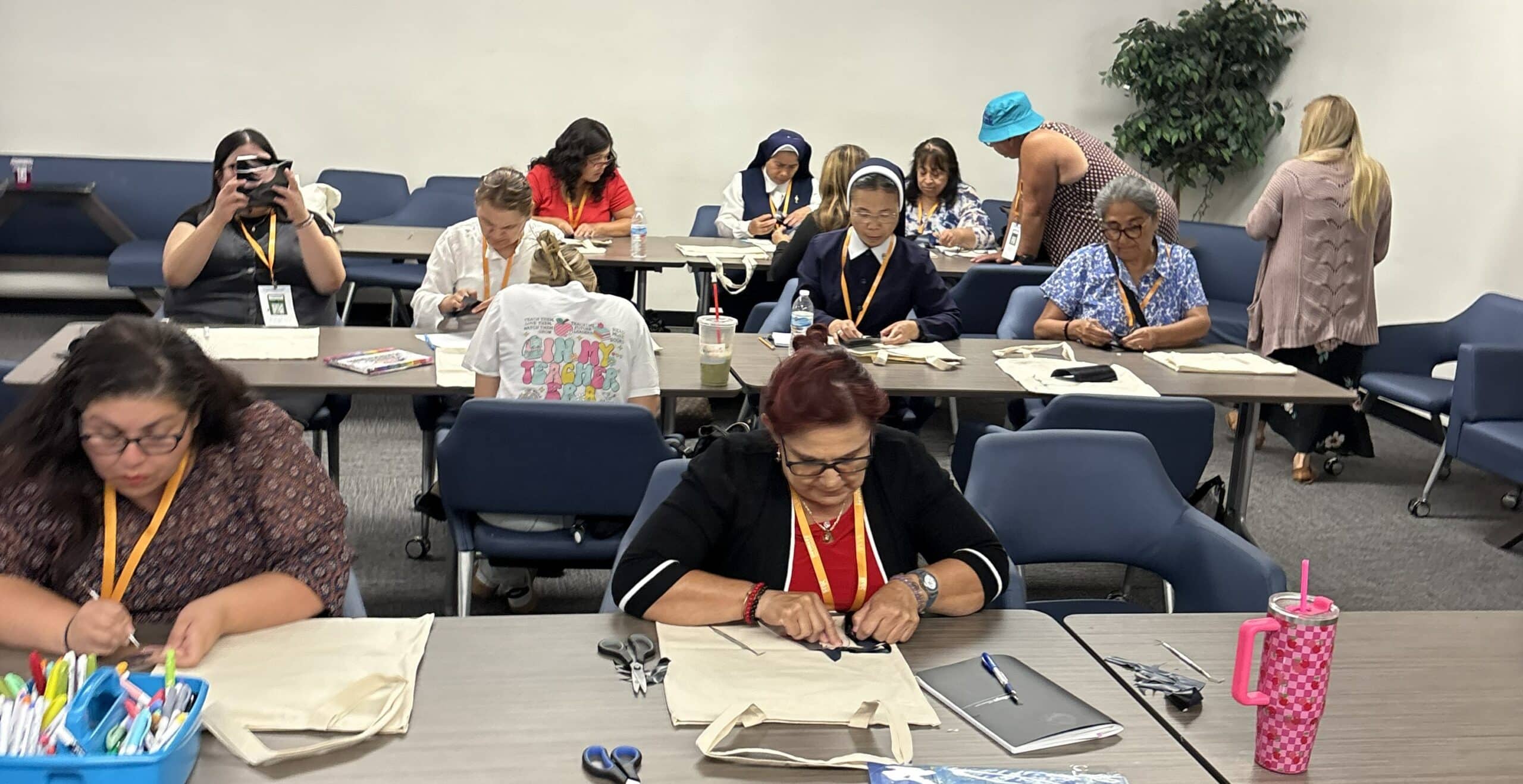 Group of adults seated at tables sewing fabric pieces in a workshop setting; craft supplies and cups on the table.