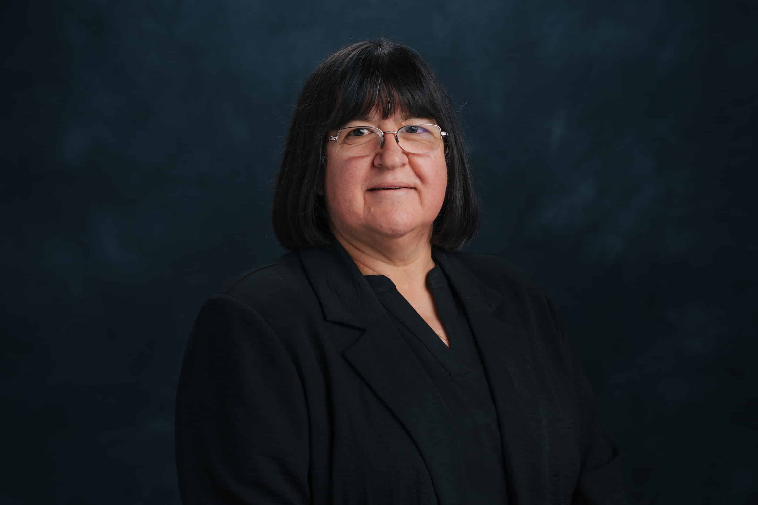 Portrait of a middle-aged woman with shoulder-length black hair and glasses, wearing a black blazer against a dark blue studio backdrop.