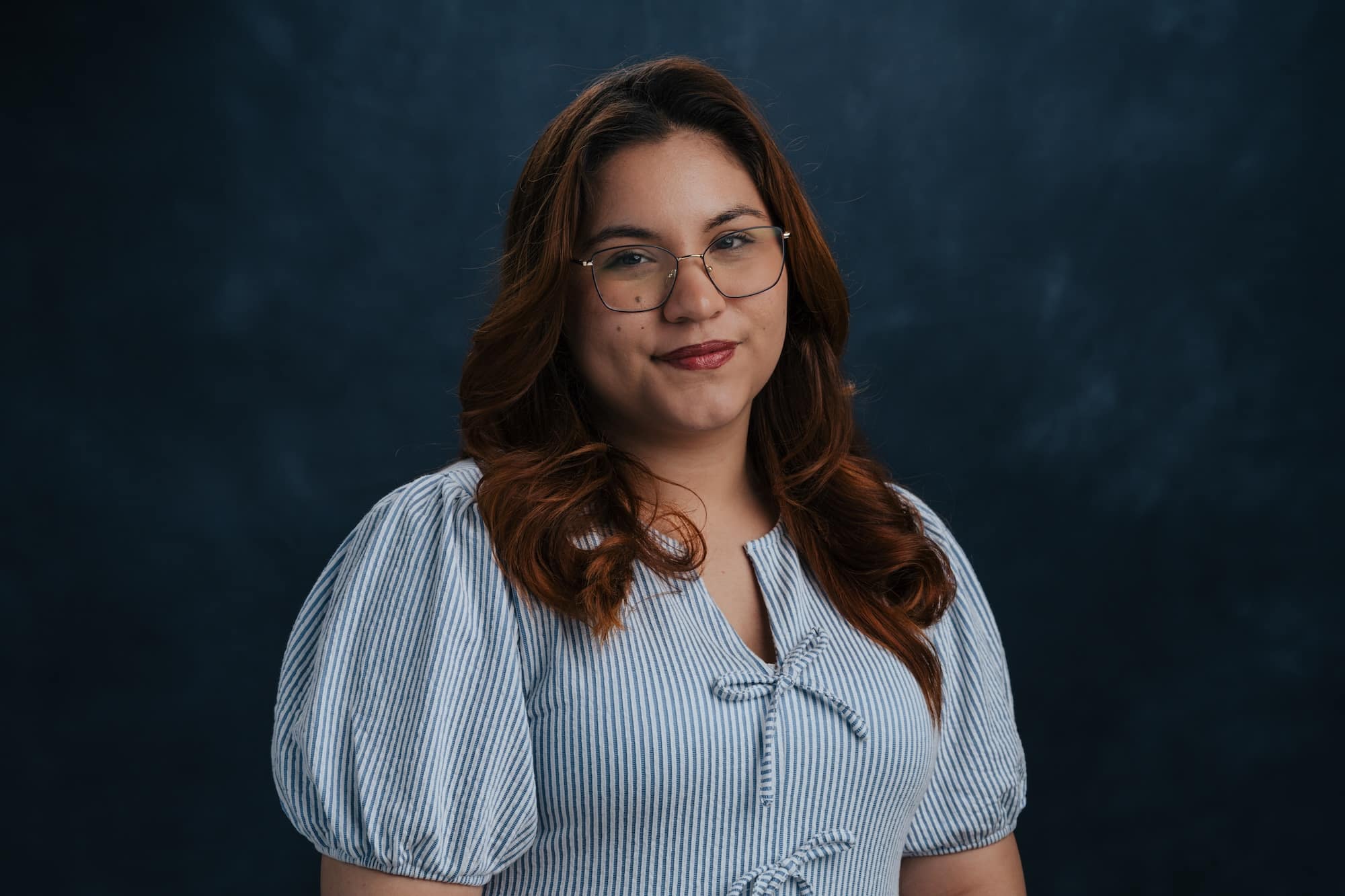 Portrait of a woman with round glasses and wavy brown hair, wearing a blue and white striped blouse, looking at the camera against a dark blue backdrop