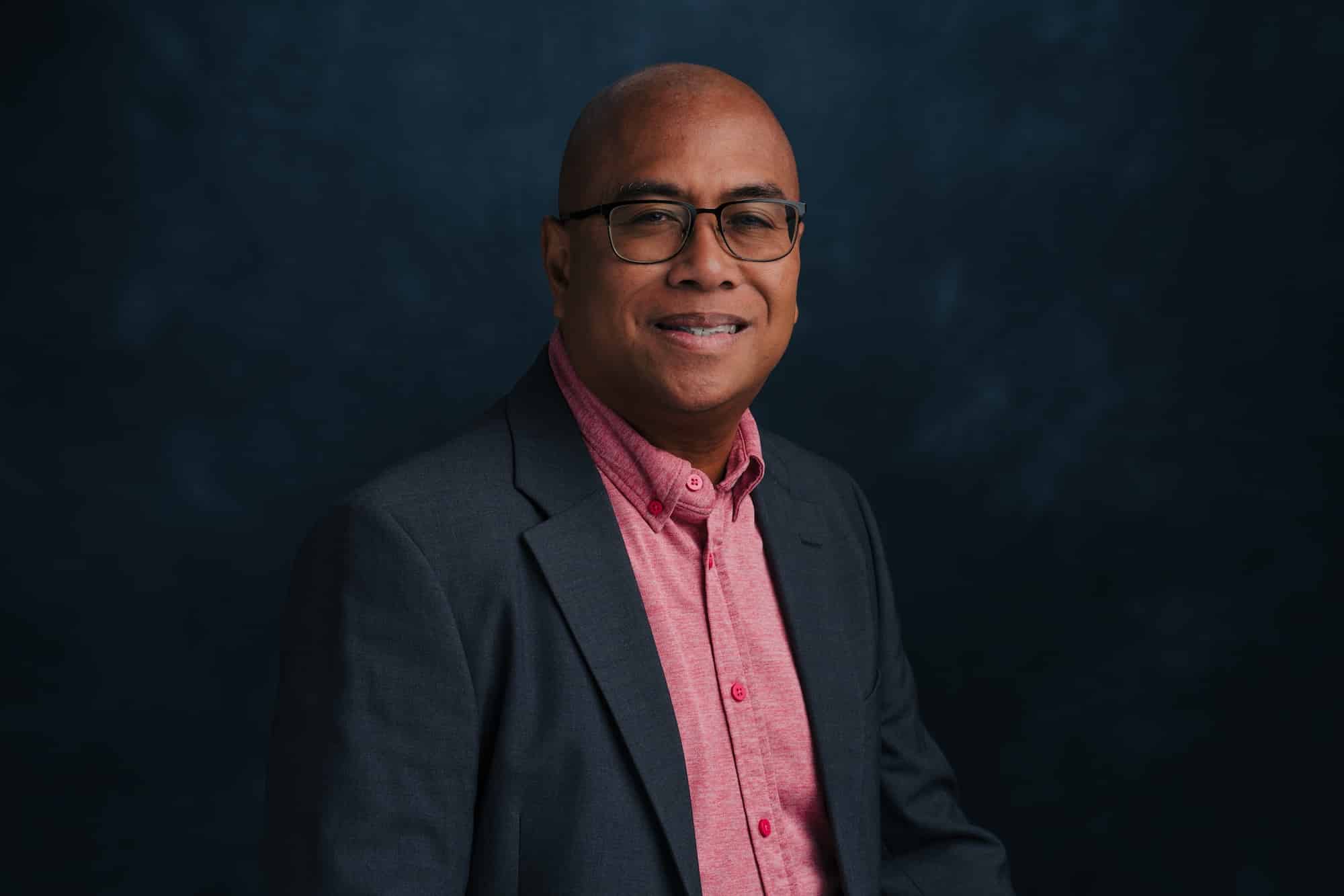 Head-and-shoulders portrait of a smiling man with glasses in a dark blazer and pink shirt, against a dark blue background.