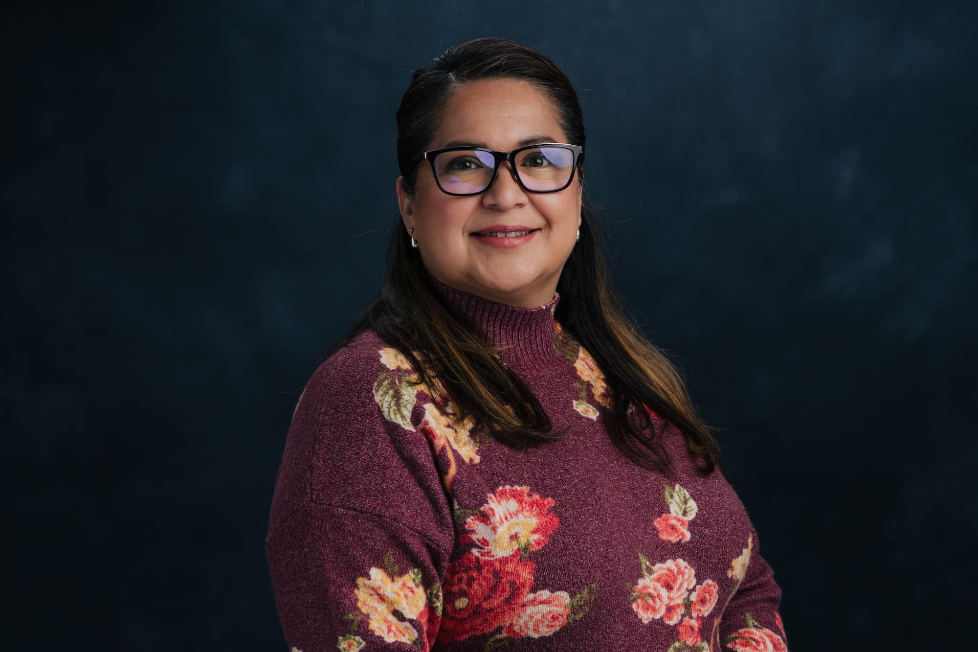 Portrait of a smiling woman with dark hair, wearing black glasses and a burgundy floral sweater against a dark blue backdrop.