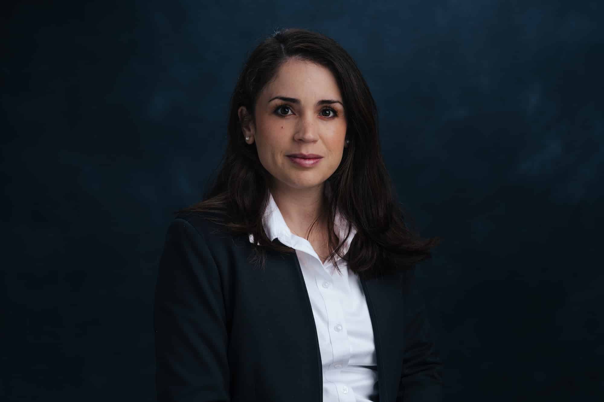 Professional woman in a dark blazer and white shirt, looking at the camera against a dark blue background.