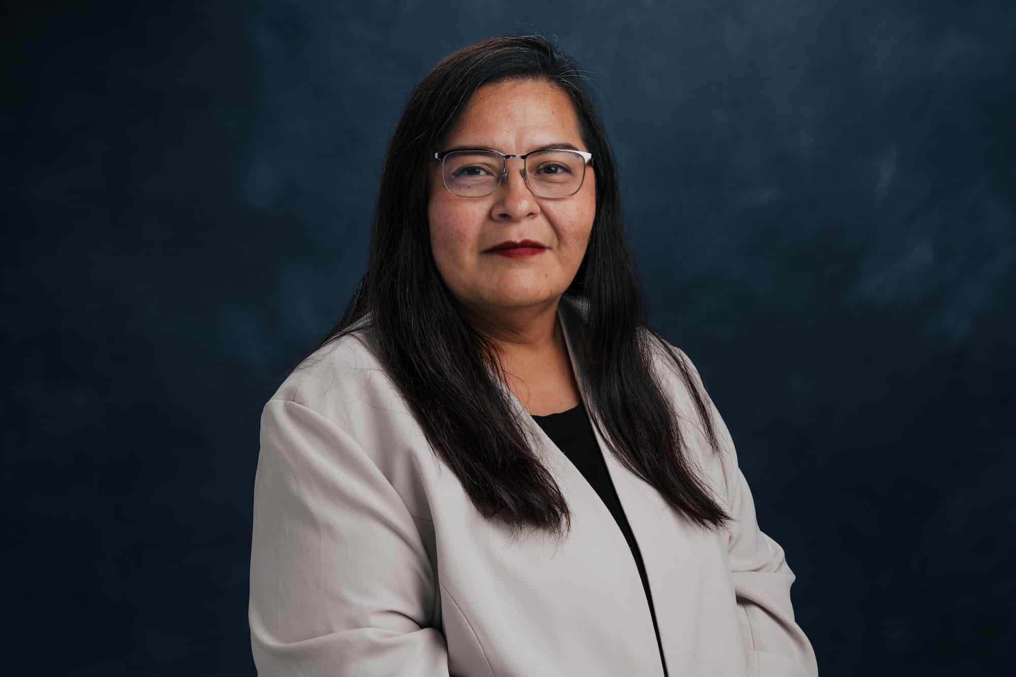 Portrait of a woman with glasses and long dark hair, wearing a light blazer against a dark blue background.