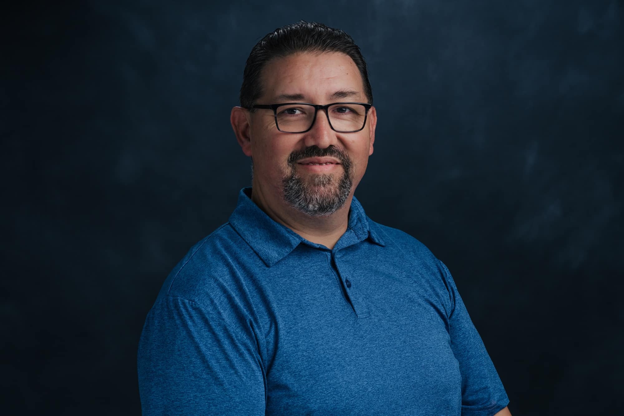 Portrait of a man with glasses, short dark hair, and a blue shirt, smiling slightly against a dark studio backdrop.