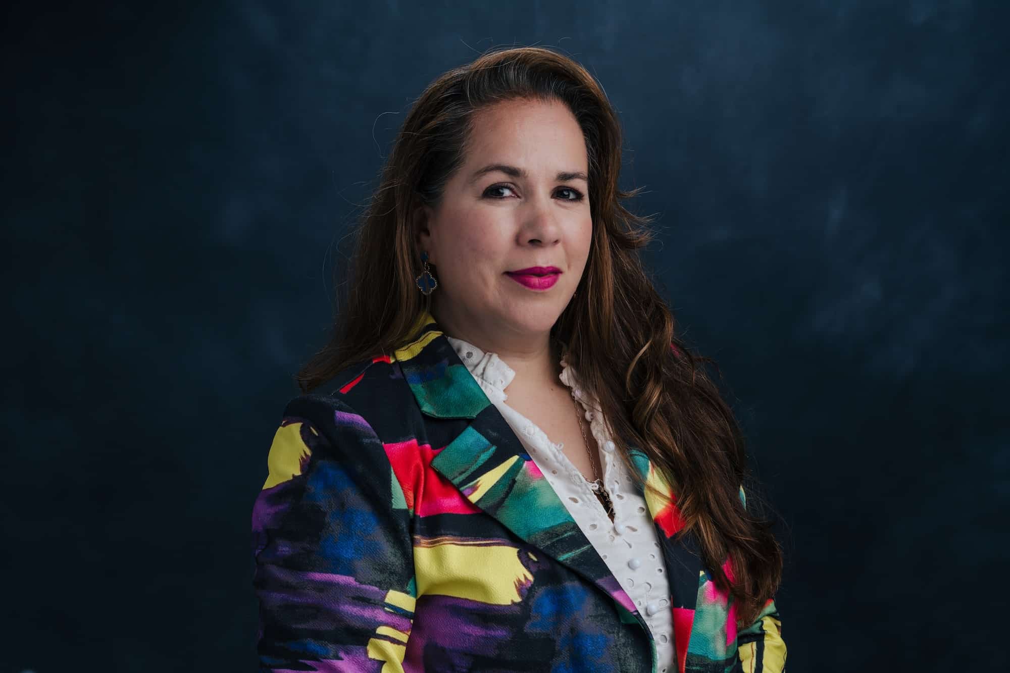 Portrait of a woman with long wavy hair wearing a multicolored blazer over a white blouse, posed against a dark blue background.