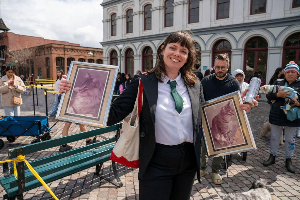 Smiling woman in a blazer and green tie holds two framed cat portraits at an outdoor event with onlookers in the background.
