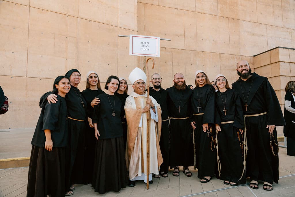 Group of nuns and monks pose with a bishop in cream robes outdoors, smiling for the photo.