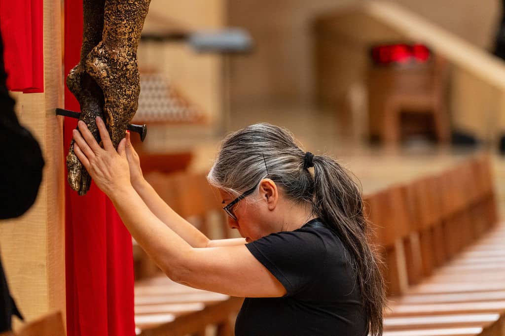 Woman in a black shirt with gray hair tied back, pressing her hands to a large statue's hands during a prayer in a church setting