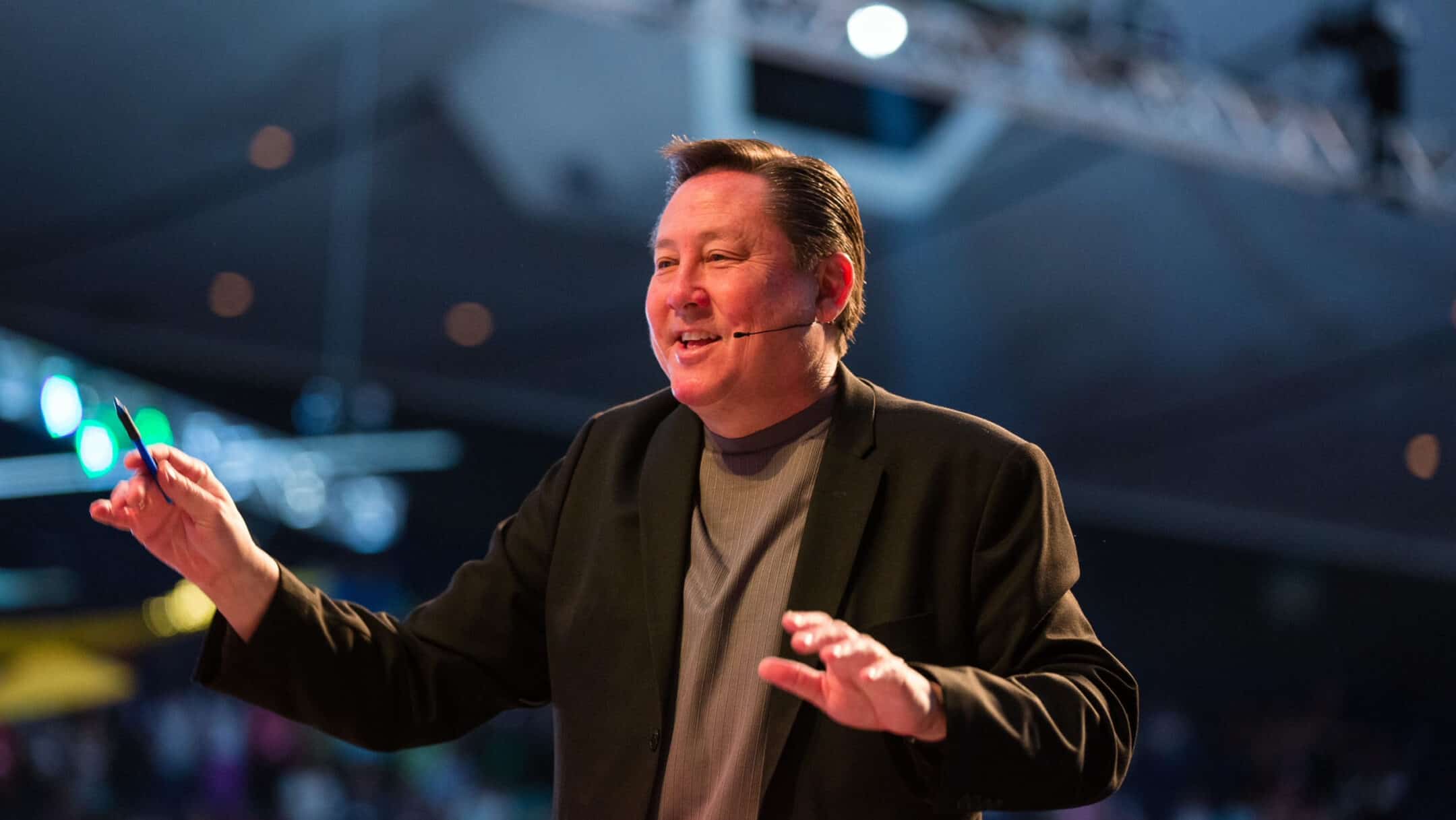 Male presenter in a blazer with a headset microphone, gesturing as he speaks on a dim stage.