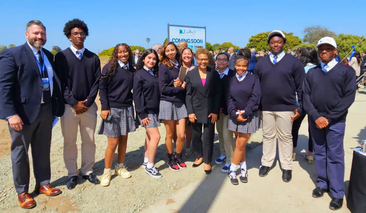 Group of students in uniform posing outdoors with a woman in a black suit; construction sign says 'COMING SOON' in the background.