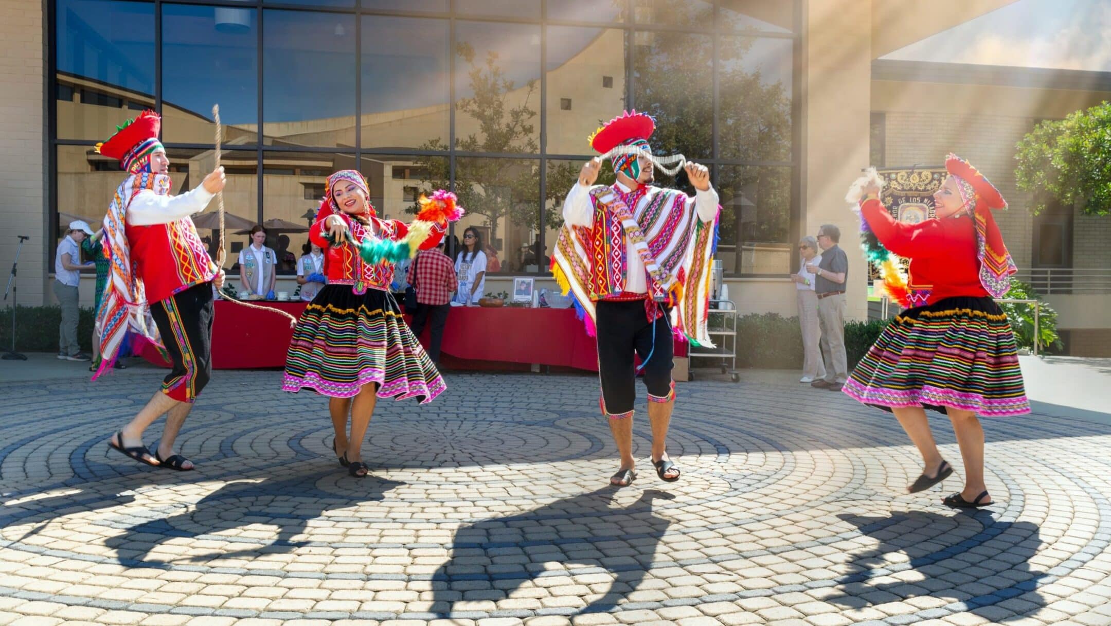Dancers in vivid, patterned folk costumes perform a lively outdoor dance on a circular stone plaza.