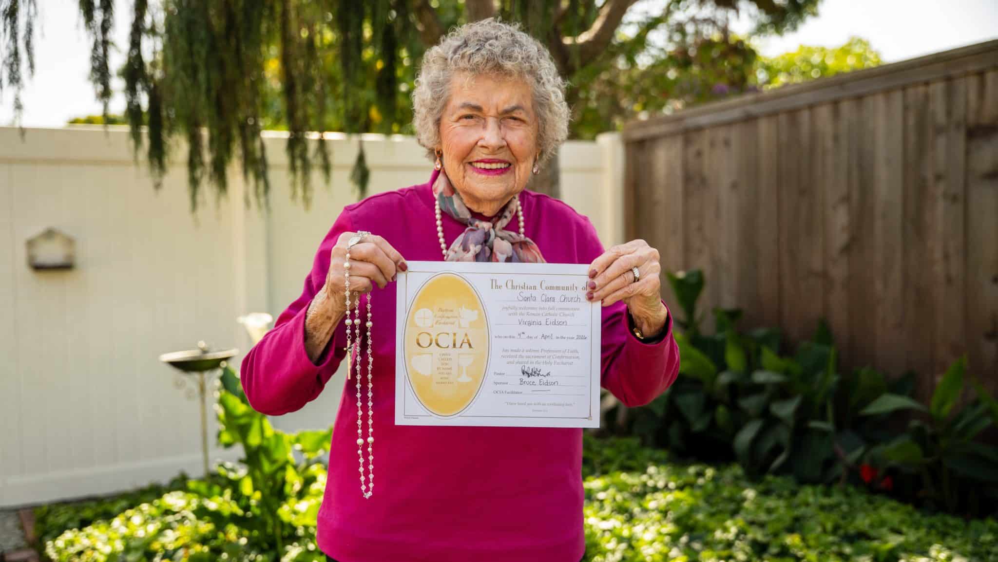 Elderly woman in a magenta top smiles while holding a certificate outdoors in a garden setting, facing the camera.