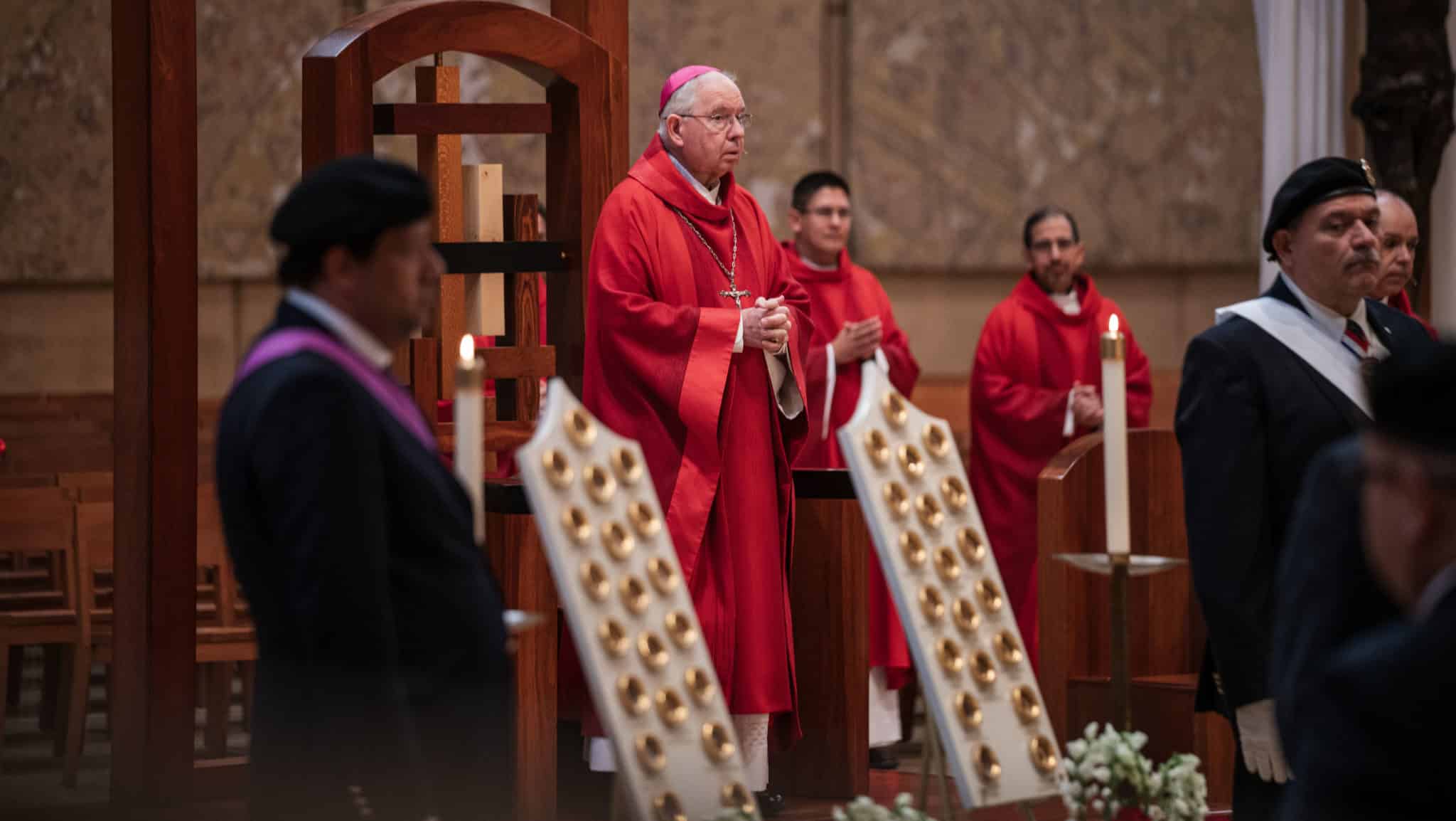 Catholic bishop in red vestments leading a service, hands folded, with congregants nearby in a church setting.