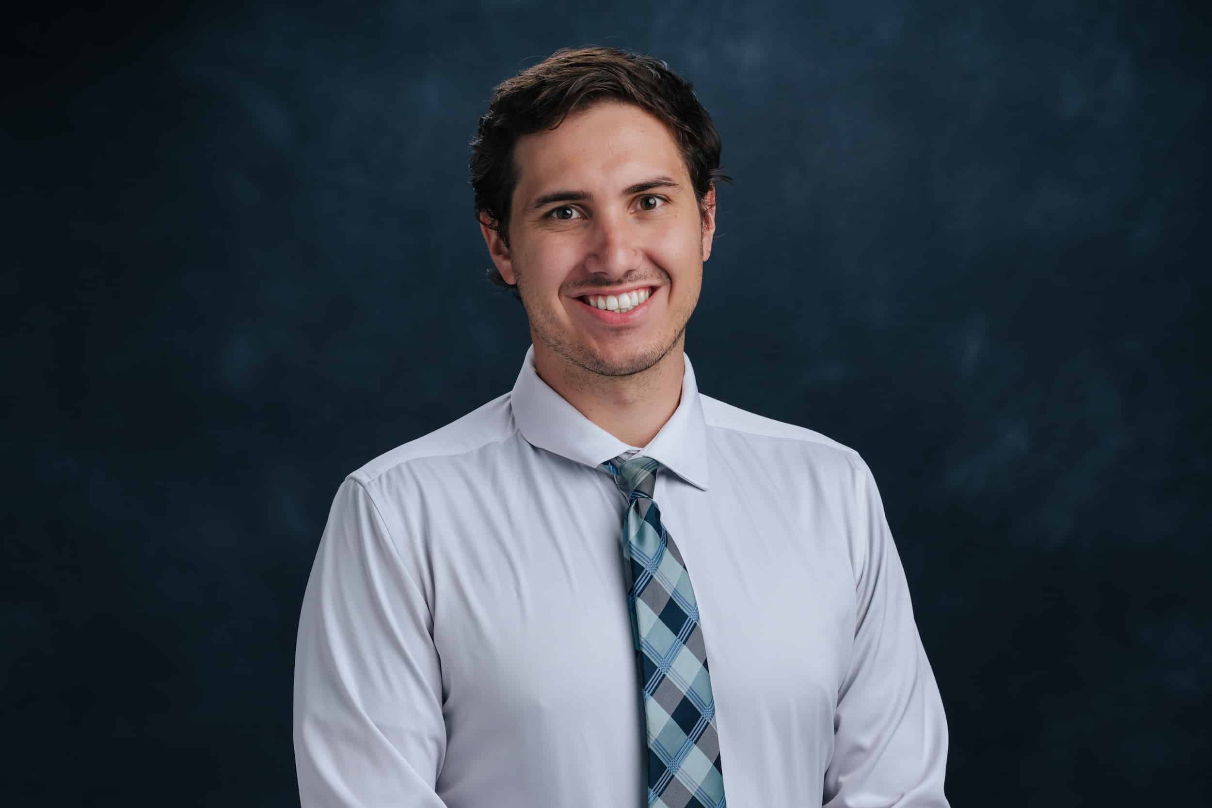 Smiling young man in a light blue shirt and plaid tie against a dark blue backdrop (portrait, head and shoulders).