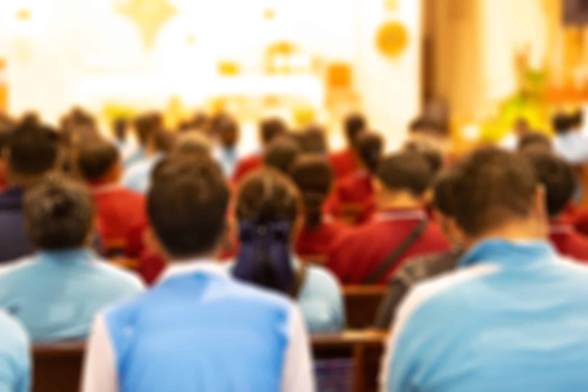 Blurred indoor conference with many people seated facing a stage or podium.