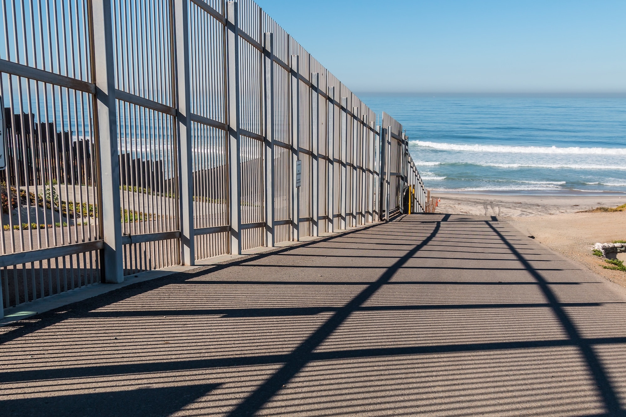 Metal fence along a sandy boardwalk leading to the ocean under a clear blue sky, with long shadows on the path.