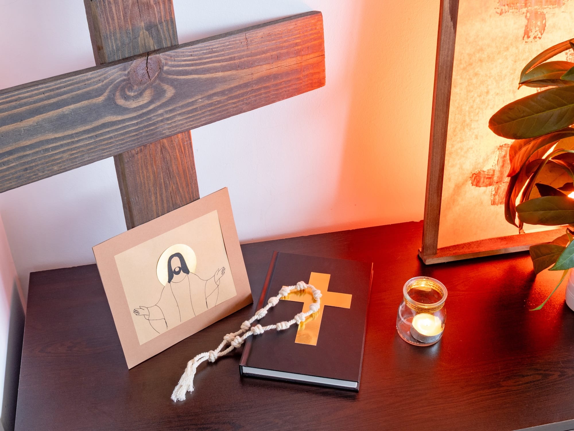Desk scene with wooden cross beams, a card showing a simple Jesus illustration, a black notebook, and a white rope on a dark surface by a lit candle.