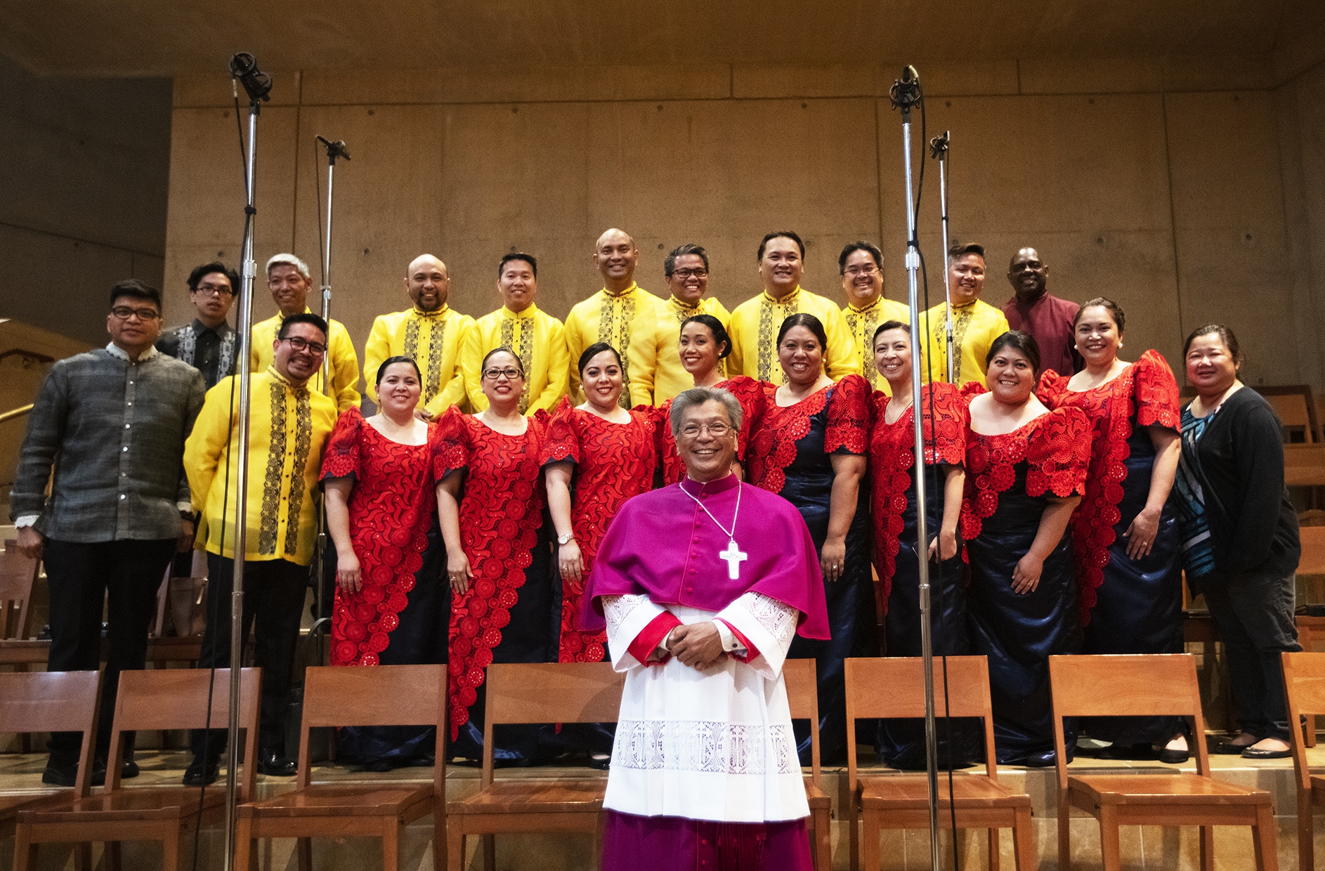 Group of singers on stage with a bishop in magenta and white robes at center, flanked by performers in red lace dresses and yellow tunics.