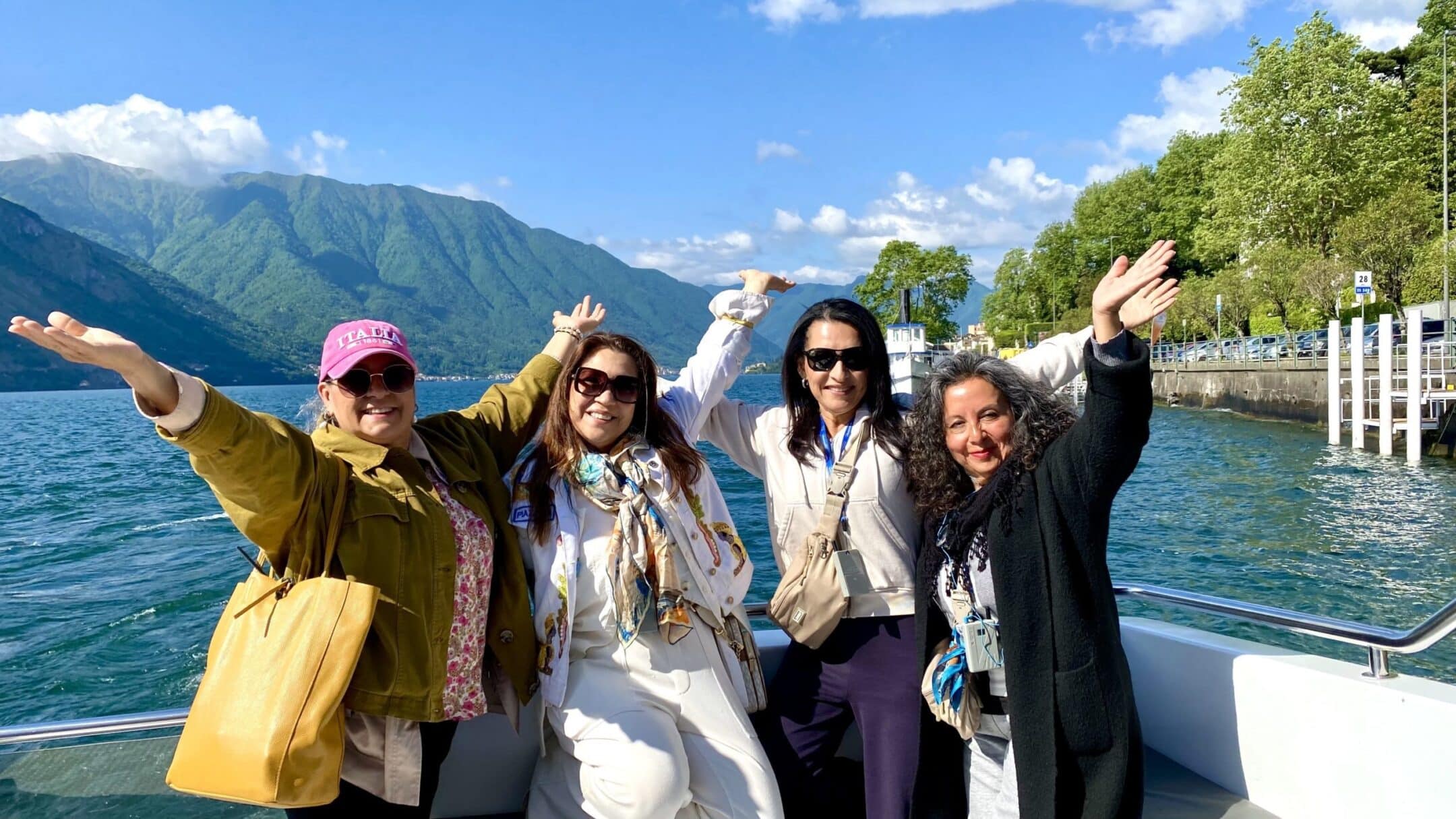Five women stand on a boat railing, arms raised, smiling against a lake and green mountains on a sunny day