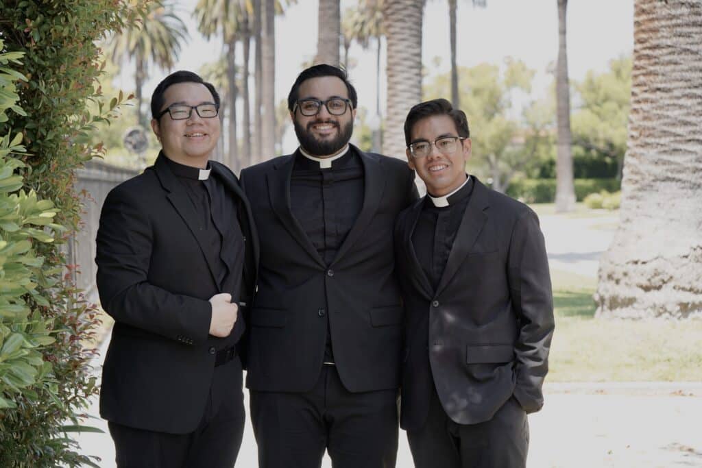 Three priests in black suits with white collars posing together outdoors, smiling among palm trees and greenery.