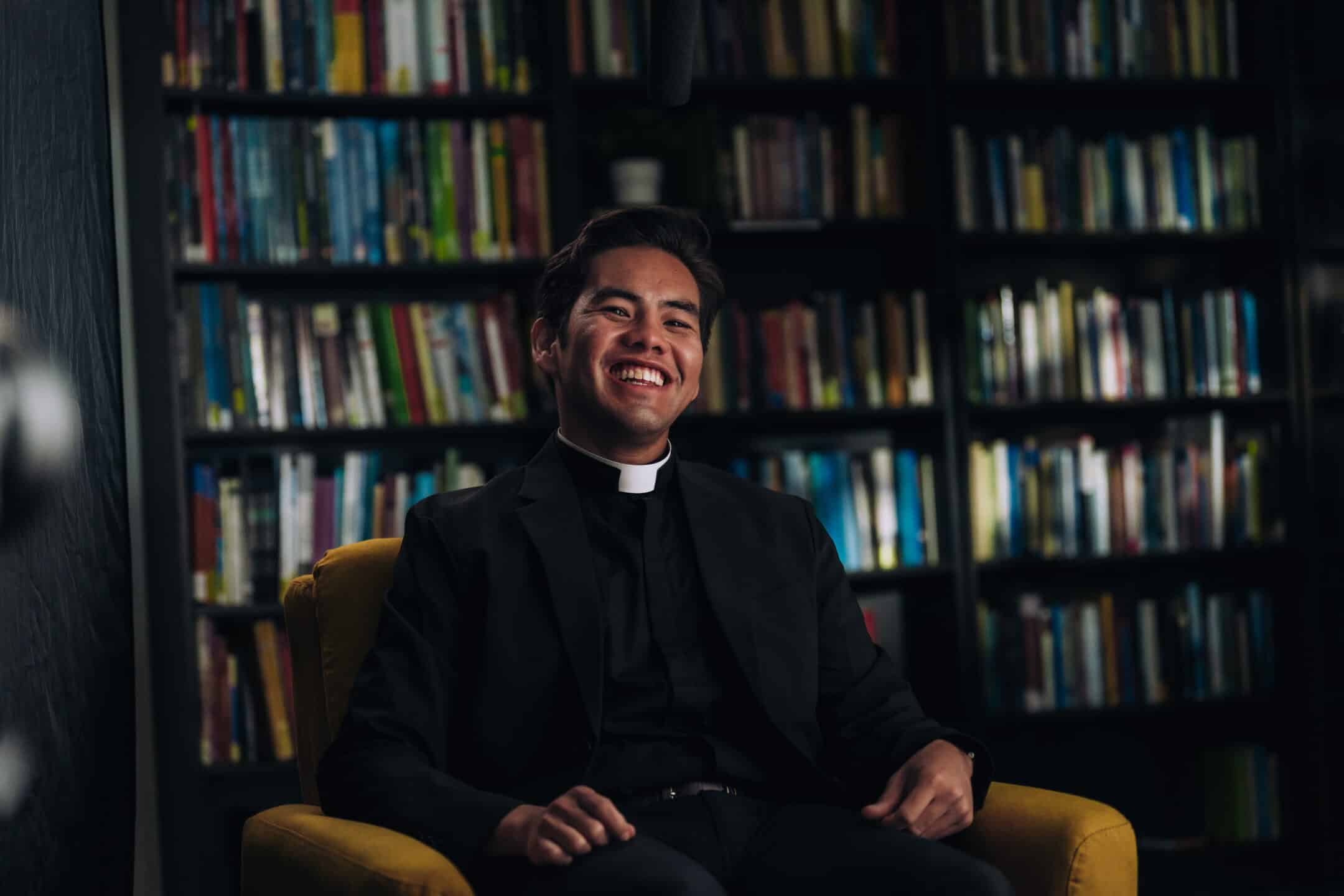 Smiling priest in a black suit and white collar sits in a yellow chair in a dimly lit library with colorful books behind him.