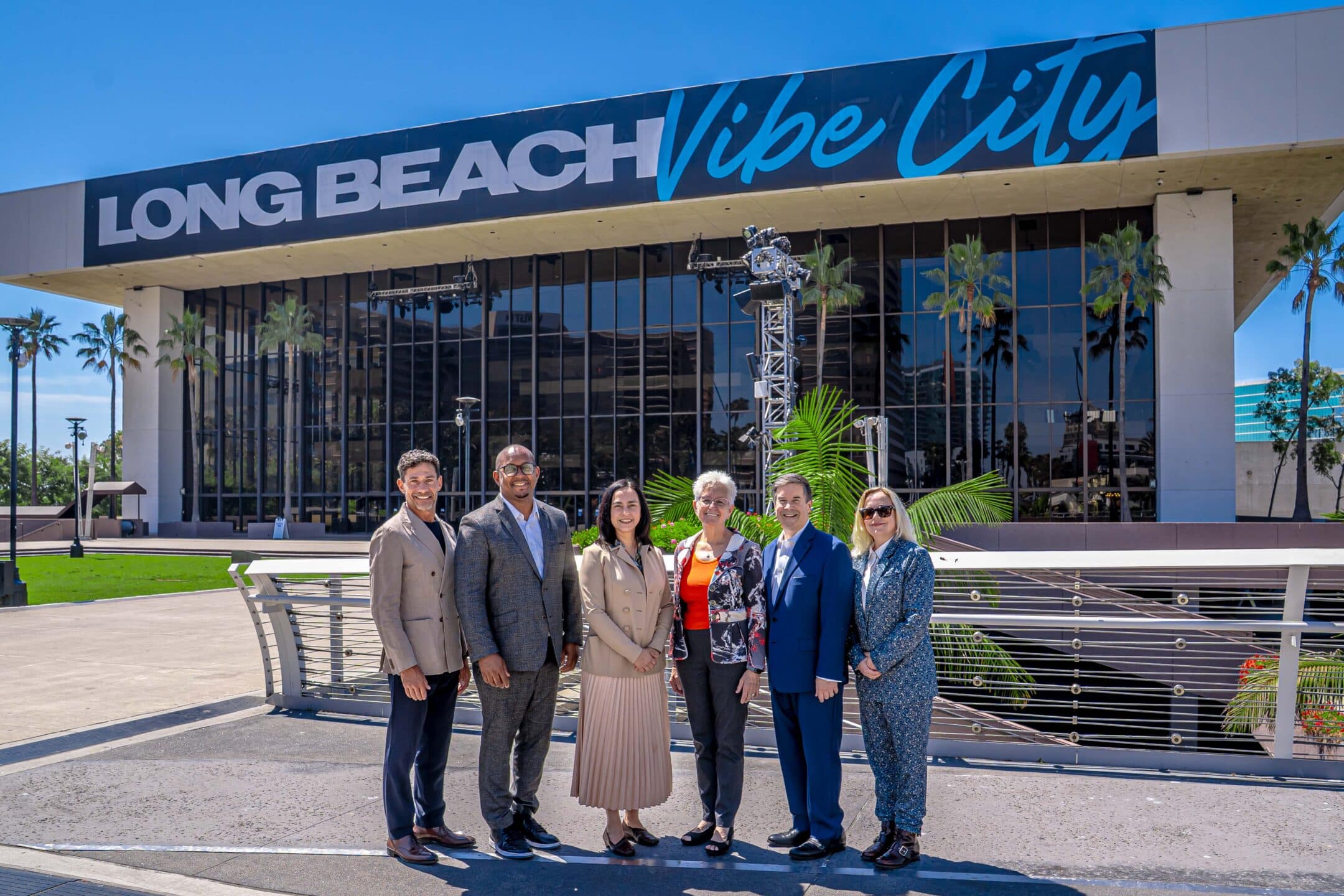 Group of seven adults in business attire posing together outside a modern glass-front building under a large 'Long Beach Vibe City' sign on a sunny day.