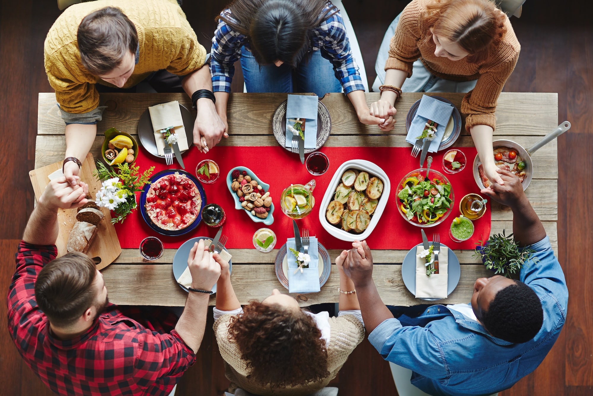 Top-down view of a group of friends around a wooden table sharing a meal, with colorful dishes and drinks arranged on a red runner.