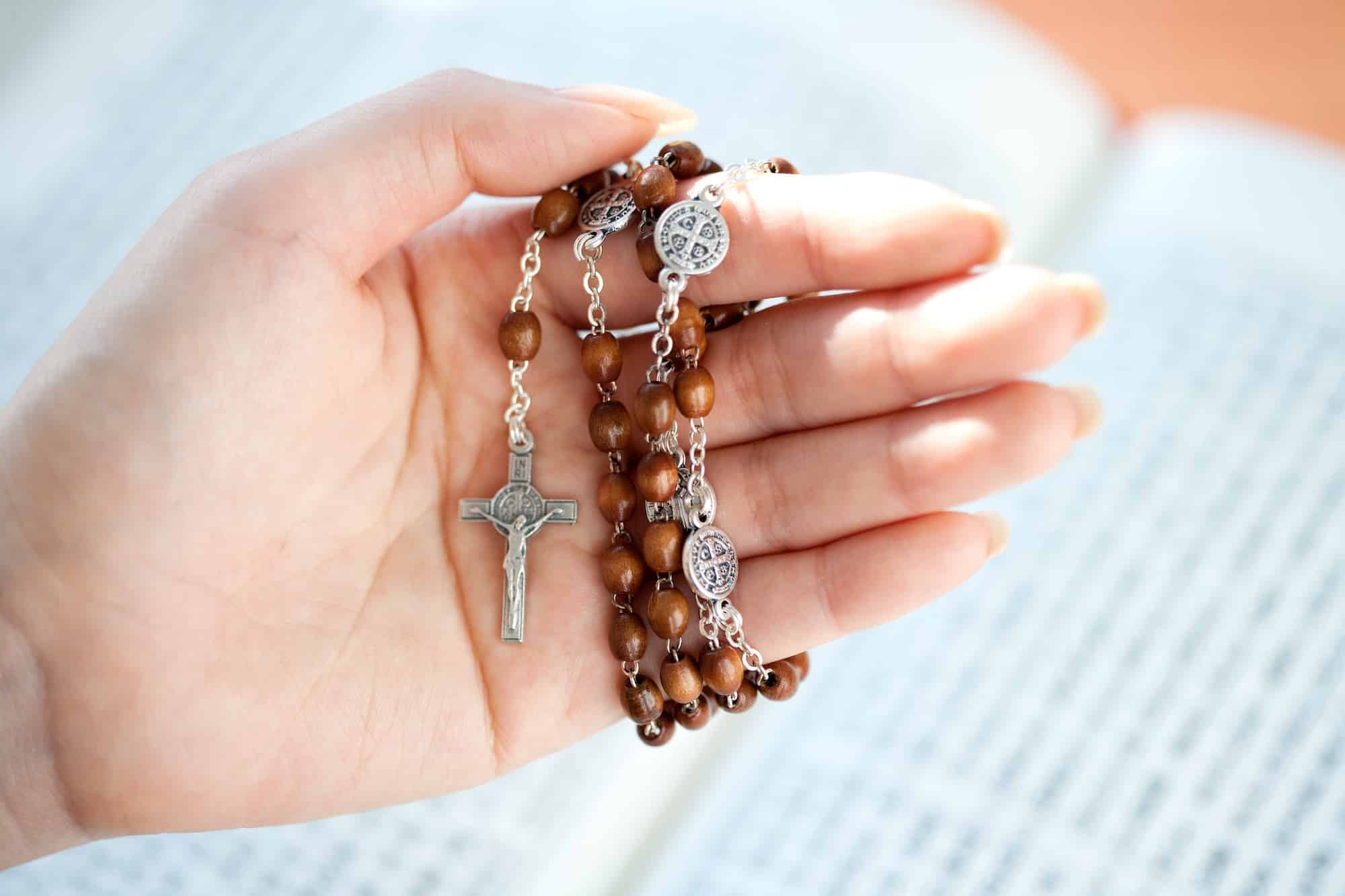 Close-up of a hand holding a brown wooden-bead rosary with a silver crucifix and round metal medals against a soft blue background.