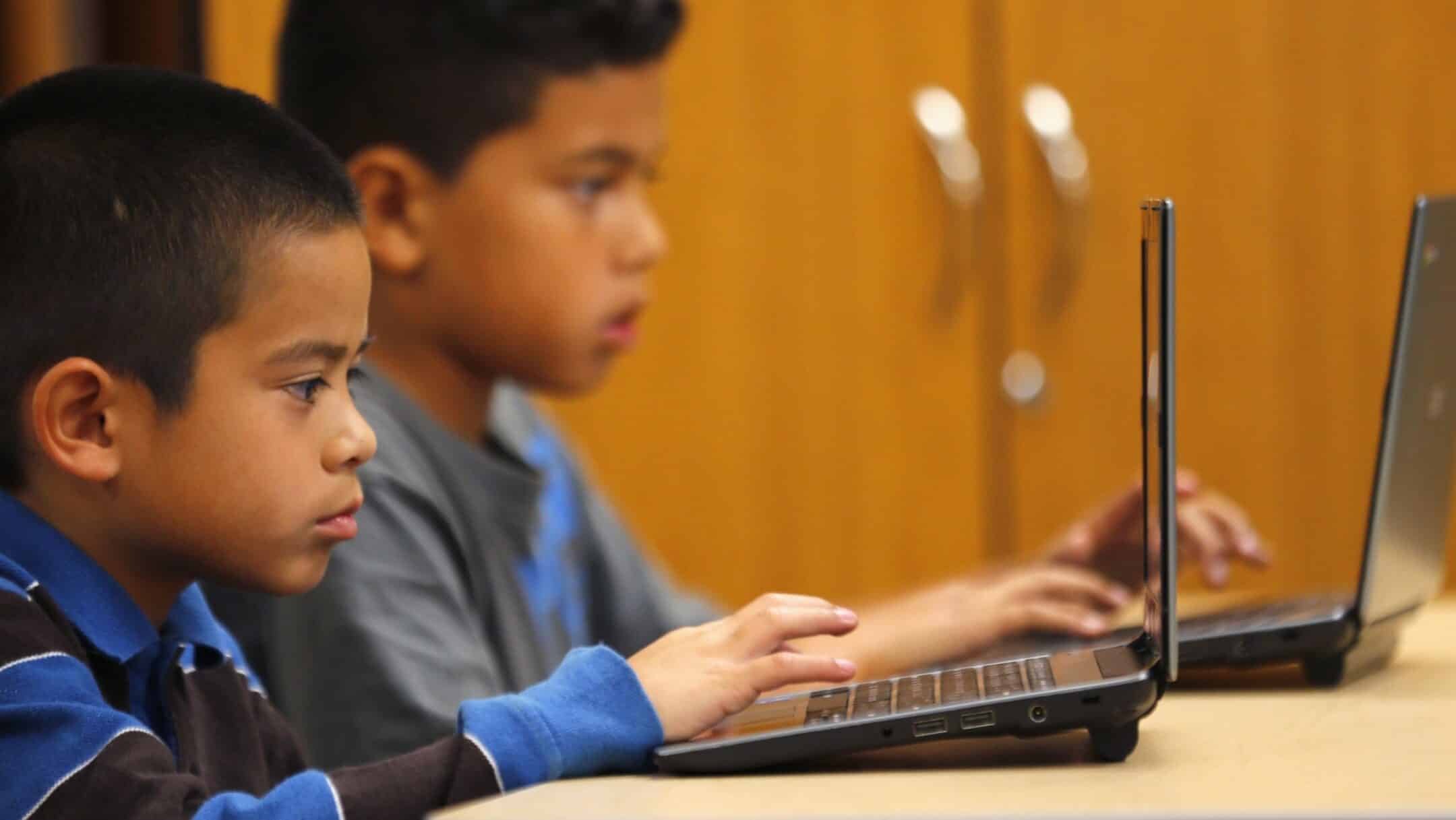 Two young students using laptops in a classroom, focused on their screens.