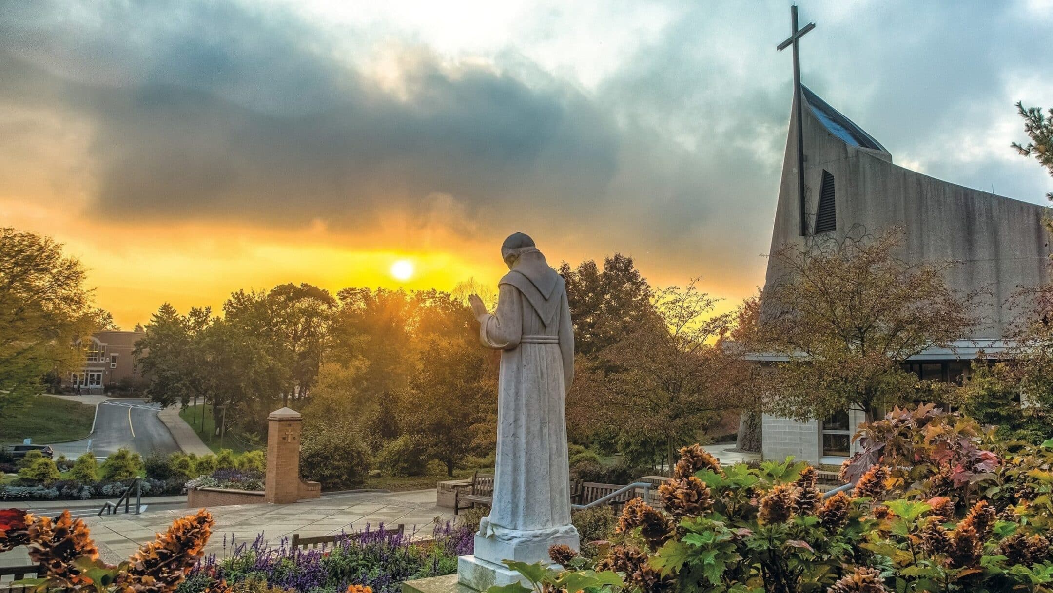 Stone statue of a robed figure in prayer in a landscaped courtyard, church with cross in the background at sunset