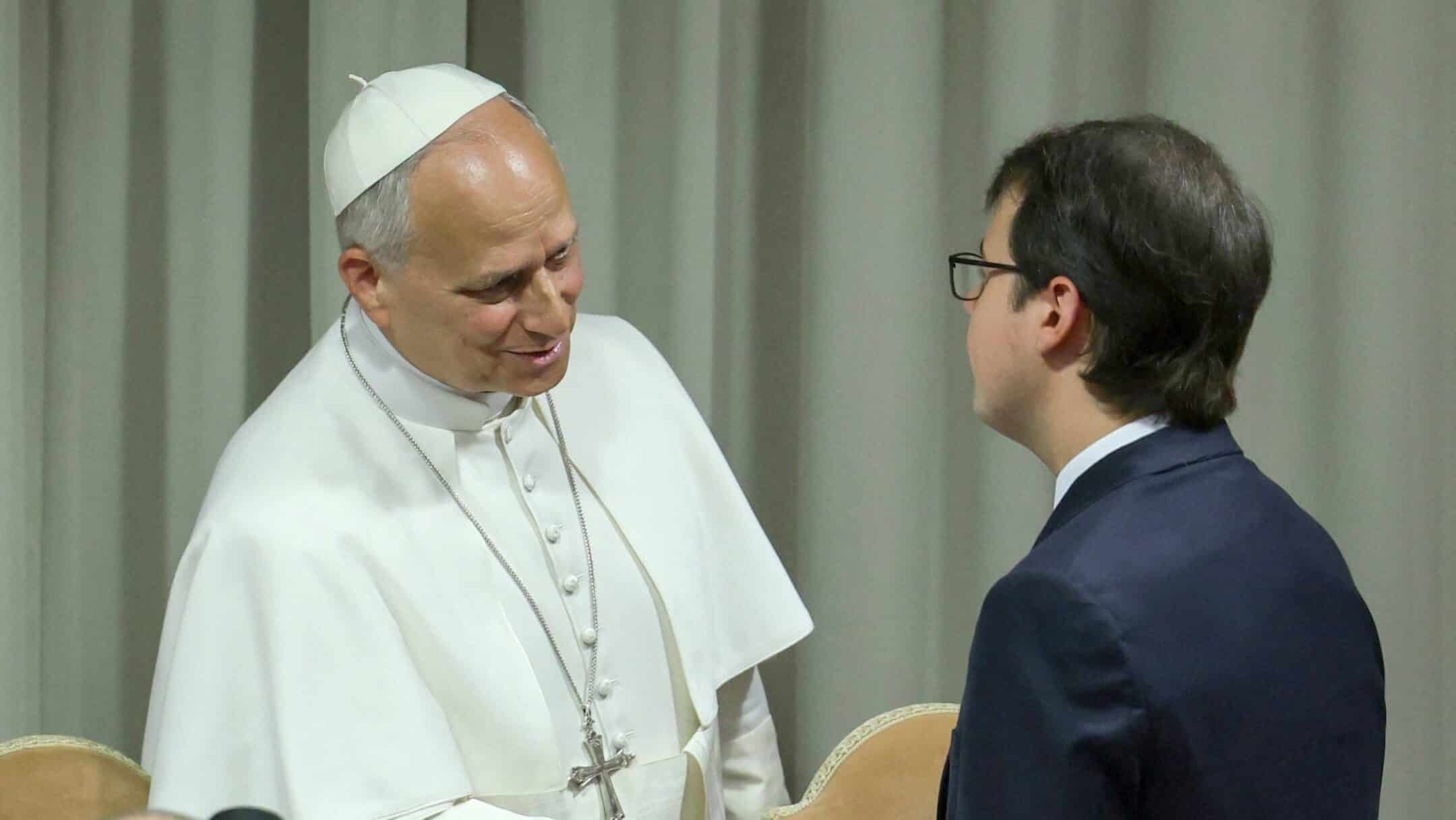 Pope Francis in white robes and a zucchetto greets a man in a dark suit indoors, smiling.