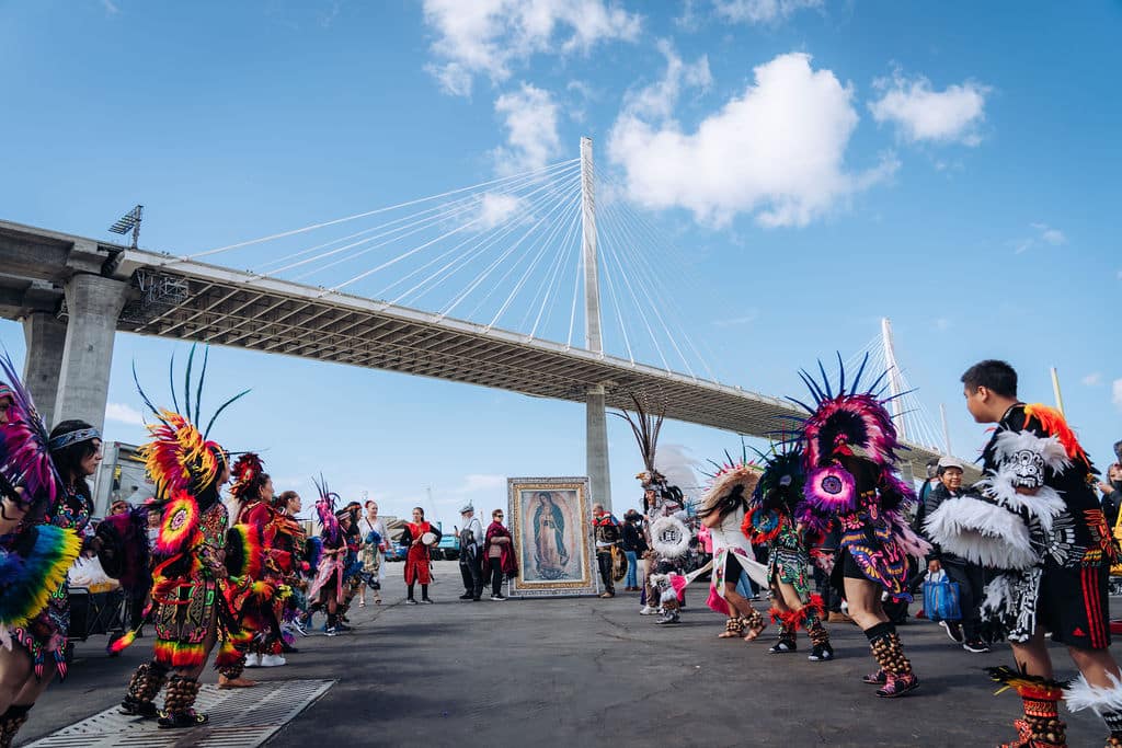 Colorful dancers in feathered costumes circle a framed religious icon under a modern bridge on a sunny day.