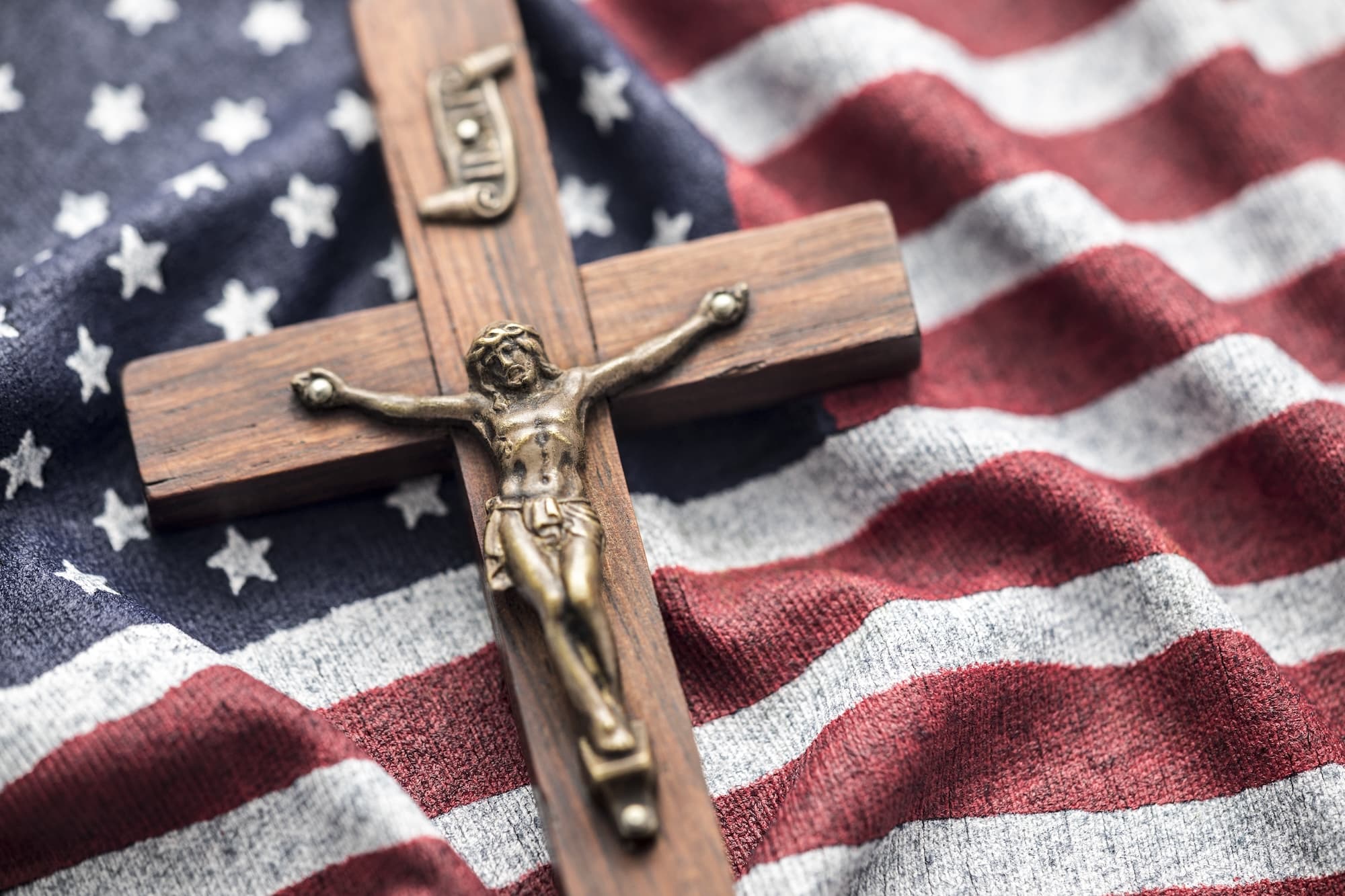 Wooden crucifix with Jesus on the cross set against a red, white, and blue American flag backdrop.