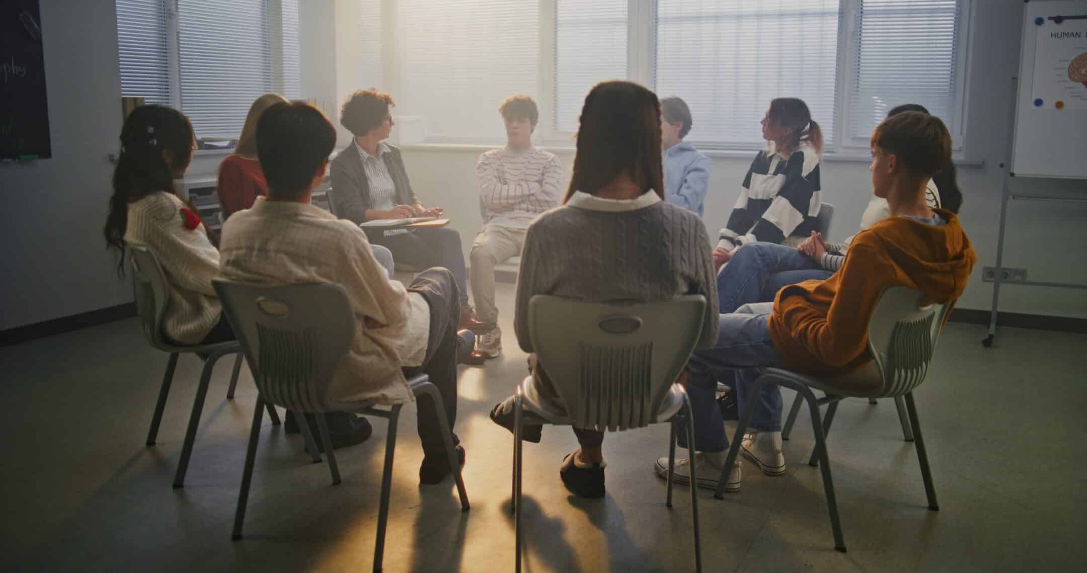 Group of students seated in a circle in a classroom, engaging in a discussion.
