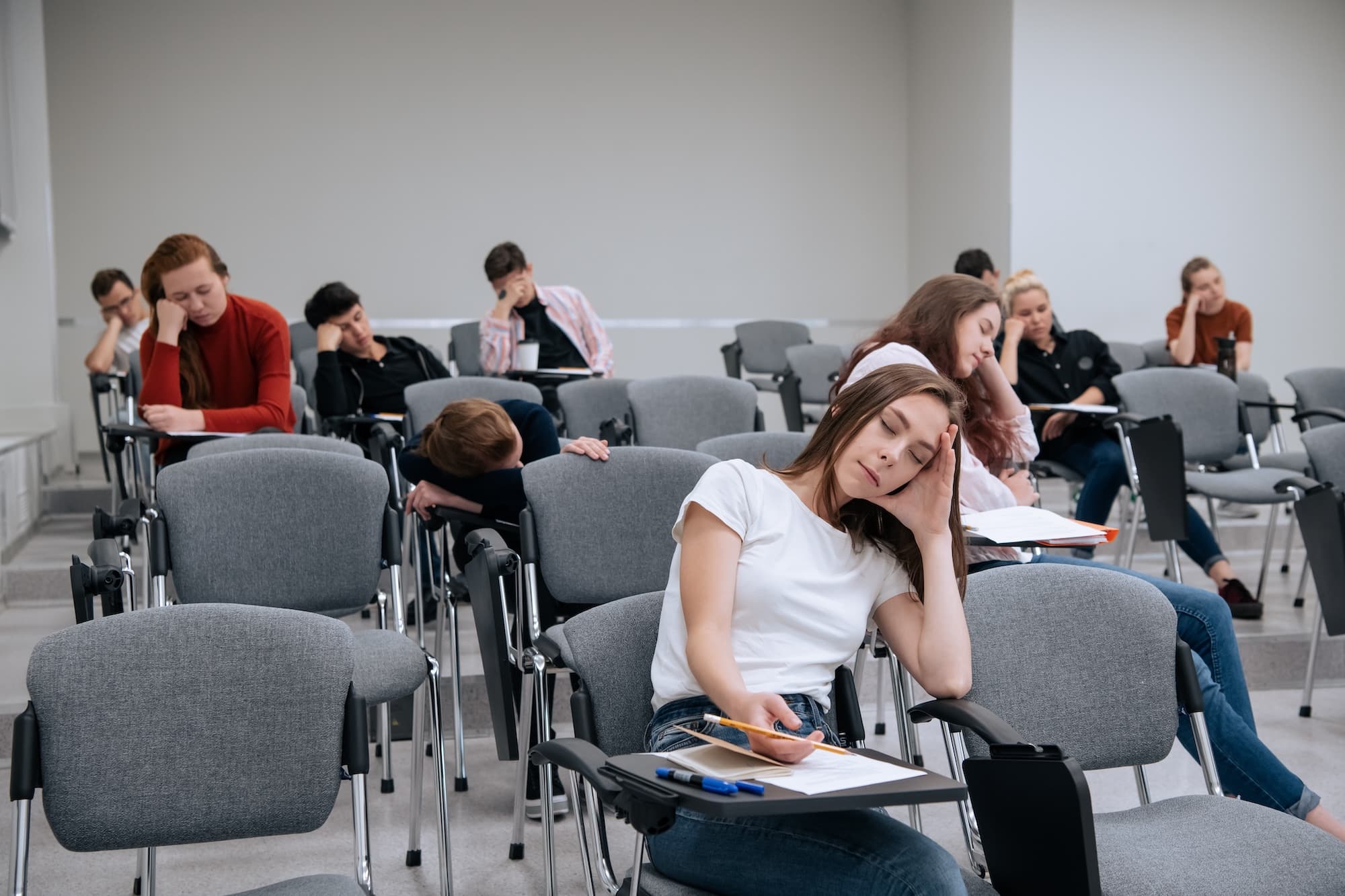 Students dozing off in a lecture hall, heads resting on desks and notebooks open.