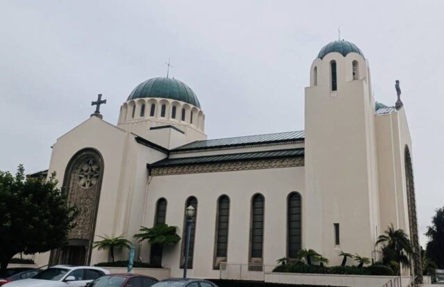 Beige church with two teal-green domes and crosses, arched windows, and parked cars in front.