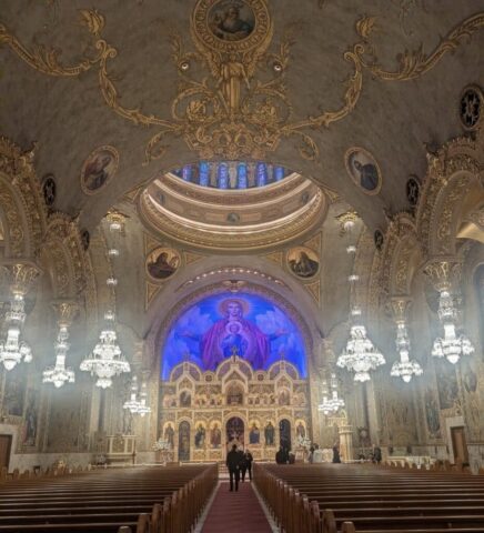 Interior of a richly decorated Orthodox church with a gilded iconostasis, chandeliers, and ornate arches overhead.