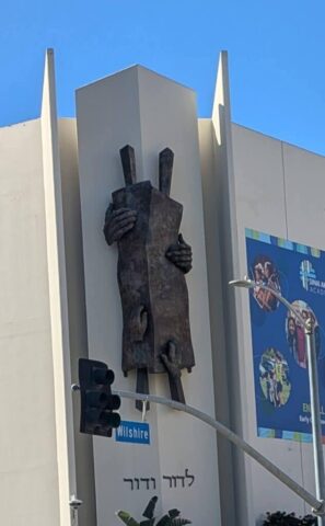 Sculpture of dark hands gripping a tall stone slab mounted on a pale building facade, under a clear blue sky; a Wilshire sign is visible nearby.