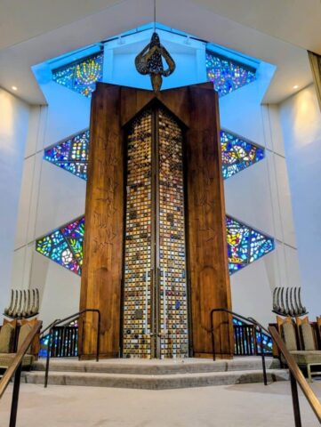 Tall wooden sculpture with a mosaic tile column framed by colorful stained-glass windows in blue-lit chapel interior, with stairs and benches nearby.