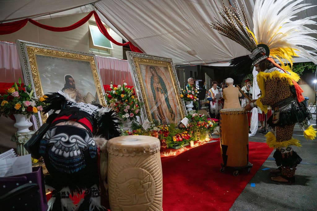 Ceremonial stage under a tent with framed religious portraits, floral arrangements, and participants in traditional dress on a red carpet.