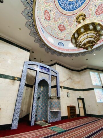 Interior of a mosque with an ornate domed ceiling of colorful patterns and Arabic calligraphy, plus a gold chandelier.