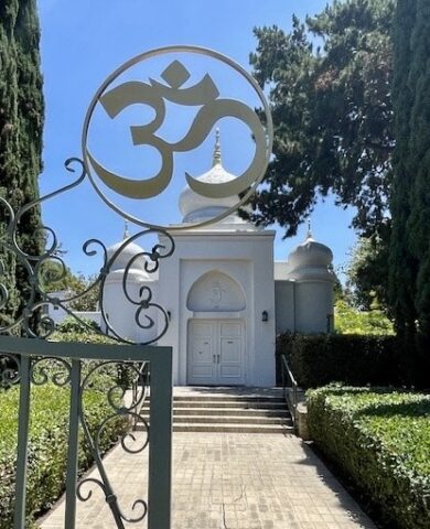 White temple entrance framed by an ornate wrought-iron gate and a large Om symbol sculpture outdoors at a sunny, gardened site