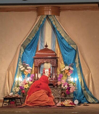 Monk in orange robes kneels before an ornate Buddhist altar adorned with blue and gold drapes, flowers and candles.