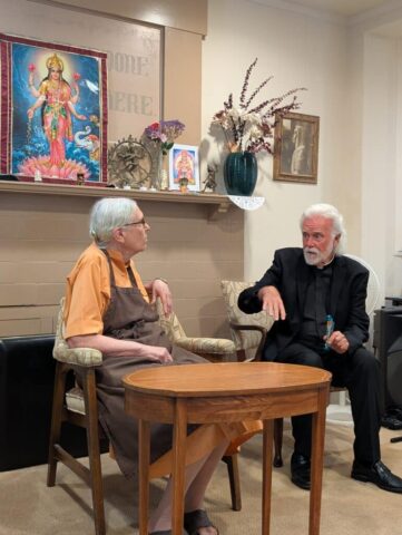 Two elderly men sit and talk in a room with a home shrine on the wall showing a Hindu goddess poster and religious artifacts behind them.